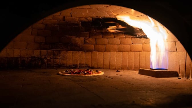 Pizza cooking by a gas powered fire in a large neapolitan brick oven